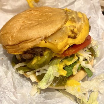 Mighty Fine Burgers Cedar Park - The Classic 1/2 lb . Hamburger with cheese - A cheeseburger features a toasted bun with melted cheese on a beef patty, topped with lettuce, tomato, pickles, and mustard. The burger is placed on crumpled white paper, with some condiments and ingredients visible around it. - Burgers Near Me - Cedar Park, Texas