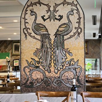 Amoo's Restaurant - Inside - A mosaic artwork depicts two peacocks facing each other, with their necks curved inward and tails intertwined, created with small tiles in various shades. The mosaic is set within an arched frame and is part of the interior decor of a restaurant, with wooden chairs and tables visible in the foreground and background. - Persian/Iranian Near Me - McLean, Virginia