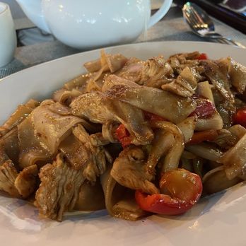 Jasmine Thai - Drunken noodles with chicken - A white oval plate holds chicken drunken noodles with sliced chicken, wide rice noodles, cherry tomatoes, and onions. The dish appears saucy and is set on a dining table with a teapot, cups, and utensils visible in the background, indicating a restaurant setting. - Thai Near Me - Lancaster, Pennsylvania