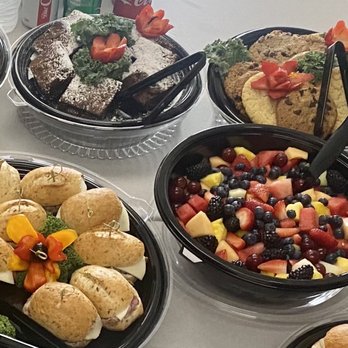 Unforgettable Edibles - Fruit salad, gourmet sandwiches, brownies and cookies. - The image shows a selection of food items on a table, including a fruit salad with berries, melons, and strawberries, along with assorted desserts such as chocolate cake and brownies. There are also savory sandwiches with toppings and garnishes, presented in black serving trays. Tongs are placed in the dishes for serving, and the table appears to be set for a buffet or catering event. - Caterers Near Me - Chicago, Illinois