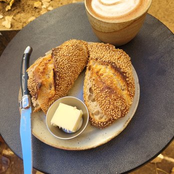 Bread Shop - My favorite Sat morning snack/ breakfast - A breakfast plate features toasted bread with sesame seeds, a small dish of butter, a coffee with foam art, and a butter knife on a black table. - Bakeries Near Me - Santa Fe, New Mexico