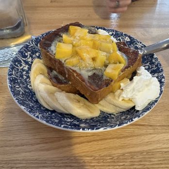 Cebu Chicago - French toast - A serving of French toast topped with fresh mango chunks, served on a patterned blue and white plate. The dish includes banana slices and a dollop of whipped cream on the side. A person’s hand and a glass of beverage are visible in the background, indicating a dining setting. - Filipino Near Me - Chicago, Illinois