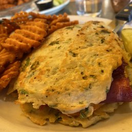 Boxty Reuben Sandwich - A boxty Reuben sandwich featuring a potato pancake wrap filled with corned beef, sauerkraut, and melted cheese, accompanied by waffle fries and a pickle spear on a plate. The dish is served in a casual restaurant setting with other dishes and drinks visible in the background.