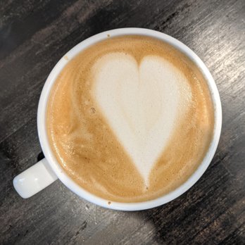 Northblock Coffee - Coffee - A white ceramic coffee cup contains a hot beverage with foam art in the shape of a heart on top. The coffee appears to be a latte or cappuccino with a light brown color, and the foam art is created with steamed milk. The cup is positioned on a dark wooden surface, viewed from above, with the handle on the left side. - Coffee & Tea Near Me - Winchester, Indiana