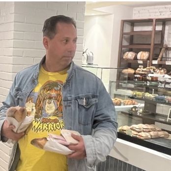 Nouvelle Maison - A man in a denim jacket and yellow graphic T-shirt holds a small puppy and some wrapped food items inside a bakery or deli. - Grocery Near Me - Winchester, Massachusetts