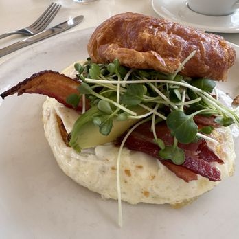 Dolina Santa Fe - Breakfast croissant - A breakfast plate featuring a fried egg topped with bacon, sliced avocado, microgreens, and a croissant, served on a white plate. - Cafes Near Me - Santa Fe, New Mexico