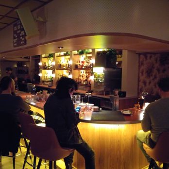 Double Fun - The image shows a bar interior with multiple patrons seated around a curved wooden counter. The bar is dimly lit, with warm lighting highlighting the liquor bottles on shelves behind the bar. Several people are engaged with their phones or talking, and a bartender is visible in the background. The setting appears cozy and modern, with a focus on socializing and drinking. - Cocktail Bars Near Me - Chicago, Illinois