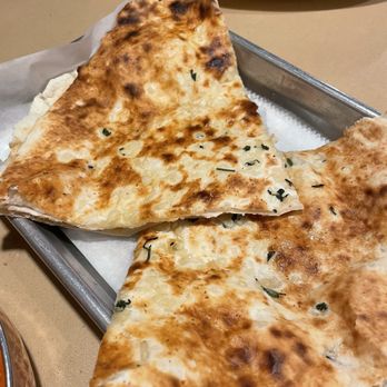 Spice Room - Garlic Naan - Two slices of garlic naan bread with herbs are placed on a metal tray, with a white plate and some utensils visible in the background. - Indian Near Me - Arvada, Colorado