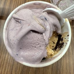 Coffee Oreo - A cup of blueberry ice cream with a creamy texture and visible blueberry pieces is shown, with a partially eaten cookie at the bottom. A white spoon is inserted into the ice cream, and a clear plastic cup with remnants of ice cream is visible in the background. The ice cream is served in a white paper cup on a wooden table surface.