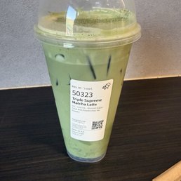 Matcha Triple supreme - A tall, green matcha latte in a clear plastic cup with a domed lid, placed on a dark table, with a paper-wrapped straw nearby.