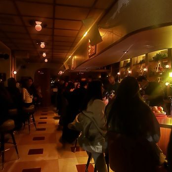 Double Fun - Bar - The image depicts a bar with a warm, dim lighting ambiance. Patrons are seated along the bar counter and at tables, engaging in conversations. The ceiling features a grid pattern with round light fixtures, and the bar area has shelves with bottles and glasses. The overall setting suggests a social environment suitable for casual gatherings. - Cocktail Bars Near Me - Chicago, Illinois
