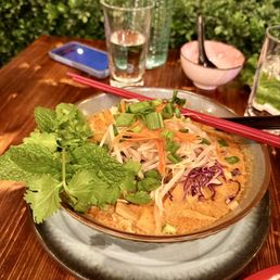 Khao Poon Gai - A bowl of Khao Poon Gai, a traditional Laotian noodle soup, is presented with bean sprouts, chopped green onions, shredded carrots, and fresh herbs like mint and cilantro. The dish is served on a ceramic plate with red chopsticks resting on top. The background includes a glass of water, a smartphone, a small bowl with a spoon, and a glass of lime water, all on a wooden table near a window with greenery outside.