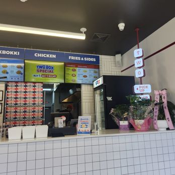 Two Box - The photo depicts the interior counter of Two Box restaurant, featuring digital menu boards listing chicken options, fries, and sides. There are flower arrangements with pink ribbons on the counter, and a black refrigerator is visible behind the counter. The setting includes a tiled counter surface and ceiling lighting, with a staff member partially visible on the left. - Korean Near Me - Englewood, New Jersey