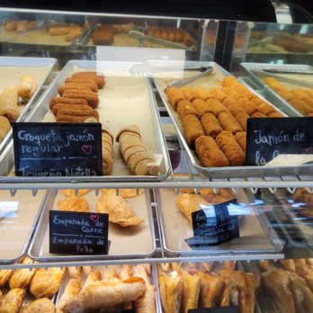 Breadman Miami - The image shows a glass display case filled with Cuban baked goods and fried foods at Breadman Miami. Items include croquetas, pastelitos, and empanadas, with handwritten labels indicating different varieties such as jamón, pollo, and carne. The case is organized with trays of food, and the reflection of the interior can be seen on the glass surface. - Bakeries Near Me - Miami, Florida