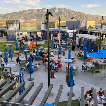 The Block - The Block.  View from Top of the Block bar. - The image shows an outdoor patio area with numerous tables and chairs, many with blue umbrellas, and some with orange and green chairs. A stage with a band performing is visible in the background, along with lighting equipment. The setting includes a grassy area, and the backdrop features a mountain range under a partly cloudy sky. The scene appears to be part of a commercial or entertainment venue with people seated and standing around. - Shopping Centers Near Me - Rio Rancho, New Mexico