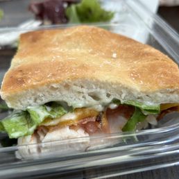 Club - A close-up of a gluten-friendly club sandwich with a toasted bread slice, lettuce, tomato, and sauce visible inside a clear plastic container. The sandwich appears partially eaten, with some greens and ingredients visible around it. The background shows additional greens and a blurred surface, indicating a casual dining or takeout setting at Urbane Cafe in Menifee, California.