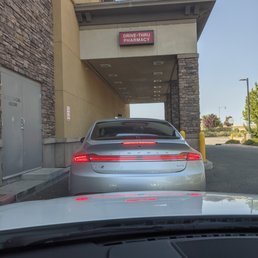 a car parked in front of a building - The image shows a silver Lincoln MKZ parked in a designated drive-thru lane at a retail store with a pharmacy. The vehicle is positioned directly in front of the store's entrance, which has a sign reading 'DRIVE-THRU PHARMACY.' The store's exterior features a combination of stone and beige wall finishes, with a covered area over the drive-thru lane. The scene is outdoors, with a clear sky and some greenery visible in the background.