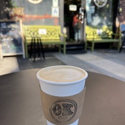 Rose Vanilla Latte - A disposable paper cup containing a latte with foam on top is placed on a dark table outside a cafe. The cup has a cardboard sleeve with a logo that reads 'OK Cafe.' In the background, the cafe's entrance features glass doors, with two green benches and a small black table visible outside. The scene is illuminated by natural light, and the cafe's signage and window decals are partially visible.