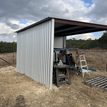Mueller - A metal shed with an open side, containing tools, a ladder, and equipment, located in a rural outdoor area with dry grass and a fence. - Building Supplies Near Me - Marble Falls, Texas