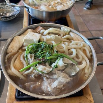 Bacogai - Lau Oxtail - Two stainless steel hot pots containing noodle dishes with vegetables, tofu, and broth are placed on a wooden table in a restaurant. The foreground hot pot has thick udon noodles, greens, tofu, and tripe, while the background hot pot appears to contain a different broth with additional ingredients. The setting includes bowls and plates, with a tiled floor and people visible in the background. - Hot Pot Near Me - Alameda, California