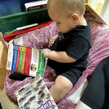 Cheeky Monkeys Fairview - Book shelf with bean bag chair to sit & read. - A young boy is seated on a pink blanket, engrossed in a colorful children's book about the world of reading, with other books nearby. - Playgrounds Near Me - Fairview, Texas