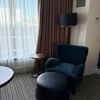The Clyde Hotel - The image shows a hotel room corner with a blue upholstered armchair and a matching ottoman placed on a striped carpet. A small round side table is positioned beside the armchair, and a tall floor lamp with a dark shade stands nearby. The window behind has sheer white curtains and brown blackout curtains, allowing natural light to enter the room. The wall includes an air vent and a thermostat, indicating a modern, well-equipped hotel setting. - Hotels Near Me - Albuquerque, New Mexico