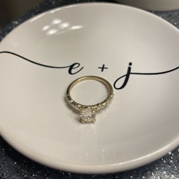 Diamond Cellar - A gold ring with a large, clear central gemstone is positioned on a white dish that has black cursive script reading 'e + j'. The dish appears to be used for jewelry display or presentation, with the ring centered on the surface. The background includes a dark, speckled surface, and the lighting highlights the ring's metallic shine and gemstone clarity. - Jewelry Near Me - Columbus, Ohio