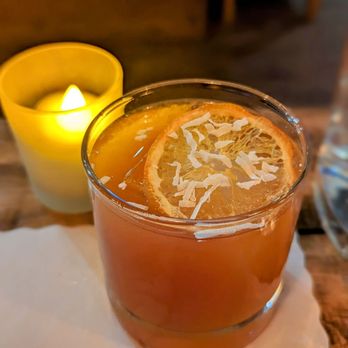 Stable DC - Rum-ination - A glass containing an orange-colored drink, garnished with a slice of lemon and sprinkled with coconut flakes, is positioned on a table. A lit yellow candle in a glass holder is nearby, creating a warm ambiance. The background includes wooden furniture and a dimly lit setting, suggesting a cozy dining environment. - Modern European Near Me - Washington, DC, District of Columbia