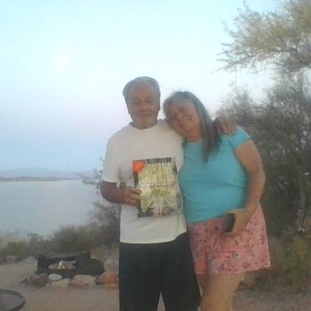 Cholla Campground - A man and woman stand close together outdoors, with the man holding a book or magazine. They are near a body of water, with desert vegetation and trees visible in the background. The scene appears to be in a natural, possibly desert, setting during daylight hours. - Campgrounds Near Me - Roosevelt, Arizona