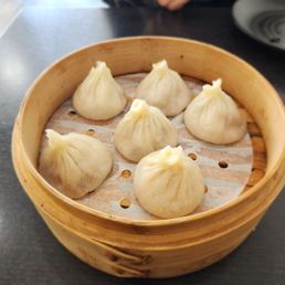 Original pork soup dumpling. - Six pork soup dumplings are placed inside a round bamboo steamer basket lined with parchment paper. The dumplings are shaped with pleated tops and are arranged in a circular pattern. The basket is on a dark-colored table, with a black plate and a white dish partially visible in the background. The setting appears to be in a restaurant, and the caption indicates they are original pork soup dumplings.