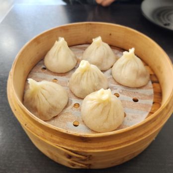 Super Bao - Original pork soup dumpling. - Six pork soup dumplings are placed inside a round bamboo steamer basket lined with parchment paper. The dumplings are shaped with pleated tops and are arranged in a circular pattern. The basket is on a dark-colored table, with a black plate and a white dish partially visible in the background. The setting appears to be in a restaurant, and the caption indicates they are original pork soup dumplings. - Dim Sum Near Me - Mountain View, California