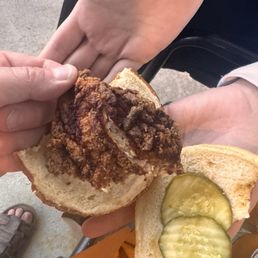 $17 chicken sandwich. This went in the garbage right after the pic was taken. - A fried chicken sandwich with pickles and bread is being held by a person, with another person's hand visible in the background.