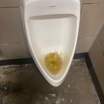 Circle K - a dirty urinal in a public restroom - A white urinal with visible yellowish stains and discoloration at the bottom, mounted on a tiled wall in a restroom. The surrounding area includes a tiled wall and a concrete floor with some dirt and stains. The urinal has a flush valve at the top and appears to be in need of cleaning. - Convenience Stores Near Me - Gilbert, Arizona