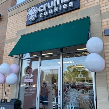 Crumbl - Naperville - Storefront - The image shows the storefront of Crumbl Cookies in Naperville, IL, with a prominent sign displaying the Crumbl logo and name. A green awning extends over the entrance, which has a glass door and large windows decorated with cookie-themed graphics. Balloons are positioned on either side of the entrance, and a small potted plant is placed near the door. Reflections of parked cars and trees are visible in the glass, and a person can be seen taking the photo inside the store. The storefront appears inviting and well-maintained. - Bakeries Near Me - Naperville, Illinois