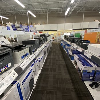 Office Depot - The image shows an aisle in Office Depot in Katy, TX, featuring multiple rows of printers and office equipment on metal shelves. The shelves are stocked with boxed printers, and promotional signage is visible above, indicating product categories. The aisle has a dark carpeted floor, and the ceiling has exposed white metal beams with fluorescent lighting. The background includes additional shelves and a clear exit door at the end of the aisle. - Office Equipment Near Me - Katy, Texas