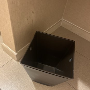 The Clyde Hotel - No trash bag ft. One of many stains all over the walls - A black plastic trash bin with a square shape and rounded edges is placed on a beige tiled floor. The bin is empty and positioned near the corner of a room, adjacent to a beige wall with a baseboard. The wall has a small stain or mark, and the floor tiles are light-colored with a textured pattern. - Hotels Near Me - Albuquerque, New Mexico