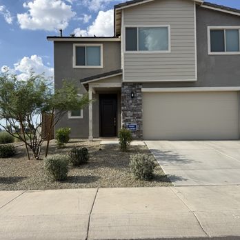 Complete Landscape Management - A two-story residential house with a landscaped front yard, including small bushes and a tree, and a closed garage door. The house has a mix of gray and beige exterior finishes. - Landscaping Near Me