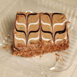 Knafeh - A rectangular layered dessert featuring a crunchy base, topped with beige icing, decorated with white and dark chocolate drizzle in a symmetrical pattern, and small green sprinkles. The dessert is placed in a clear plastic container on a light-colored surface, with some crumbs and shredded topping visible around it.
