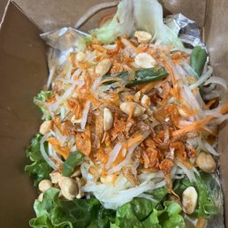 Papaya Salad - A papaya salad with shredded green papaya, carrots, and bean sprouts, topped with peanuts and crispy fried shallots. The salad is served in a brown takeout container with foil lining, and includes leafy lettuce at the bottom. The dish appears fresh and colorful, typical of Thai cuisine.