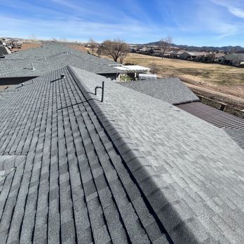 TSM Roofing - The image captures an aerial perspective of a residential neighborhood with multiple houses featuring gray shingle roofs. The roofs are aligned in a row, with some vents and pipes visible. Surrounding the neighborhood are open grassy areas, trees, and distant mountains under a partly cloudy sky. The scene emphasizes the uniformity of the roofing and the suburban setting. - Roofing Near Me - Phoenix, Arizona
