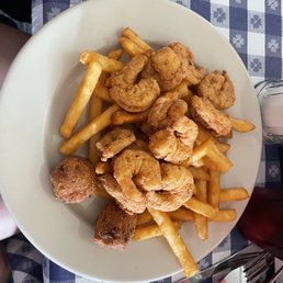 Shrimp plate - A white oval plate contains fried shrimp, French fries, and two hush puppies. The plate is set on a blue and white checkered tablecloth with a fork and knife nearby. The setting appears to be a casual dining environment.