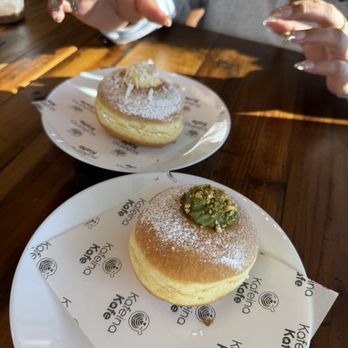 Kafeina Kafe - I would definitely recommend the bomboloni both flavors are sooo good! - Two plates of pastries, one topped with green cream and pistachios and the other with powdered sugar, are on a wooden table. A person reaches for one pastry. - Coffee & Tea Near Me - Spring, Texas