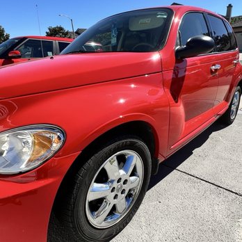 Carfection Pro - Auto body painting red in Oxnard - A red compact car is parked on a concrete surface outdoors, with a bright, clear blue sky above. The vehicle features tinted windows, black side mirrors, and alloy wheels. Part of another red vehicle is visible in the background, along with a streetlamp and some trees. The scene suggests a parking lot or driveway on a sunny day. - Body Shops Near Me - Oxnard, California