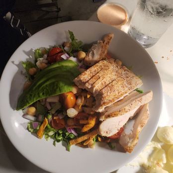 Cave Restaurant - A white plate contains sliced grilled chicken, a fresh side salad with cherry tomatoes, corn, and mixed greens, and avocado slices. The table also has a glass of water, a piece of bread, and a fork. The setting appears to be a restaurant with a casual dining atmosphere. - Venues & Event Spaces Near Me - Temecula, California