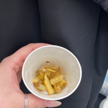 Five Guys - "Little Fries" - A person holds a small paper cup with leftover French fries, with a dark fabric surface in the background. - Burgers Near Me - Bayside, New York
