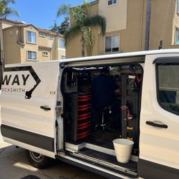 A locksmith technician is seated inside a white service van, working with tools and equipment, with residential buildings and palm trees in the background.