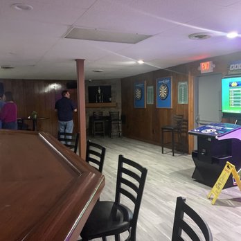 The Hideaway Tavern - a game room with a pool table - The image shows an indoor bar or recreational area with dartboards on the wall, chairs, and a gaming machine, with a few people present. - Sports Bars Near Me - Hebron, Connecticut