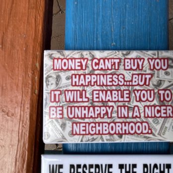 Guerrilla Graphix - A rectangular sign features a humorous quote about money and happiness, stating that money can't buy happiness but can lead to a nicer neighborhood. The sign has a background of dollar bills and is placed on a blue bench with wooden armrests, outdoors on a paved surface. - Printing Services Near Me - Albuquerque, New Mexico
