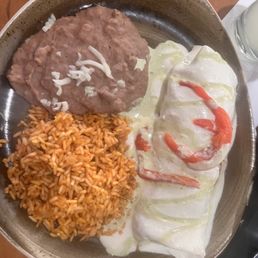 Chicken Burrito - The image shows a round plate with Mexican food items: a portion of seasoned rice, a beef dish topped with sauce and cheese, and an enchilada covered in white cheese sauce with red sauce drizzle. The plate is placed on a wooden table, with a glass of beverage visible in the background.