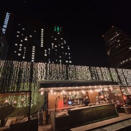View of the JW Marriott towers and the bar area. - Nighttime scene of a rooftop bar with string lights hanging above the bar area. The background features tall buildings with illuminated windows and signage, including the JW Marriott. The bar has a counter with staff preparing drinks and guests standing nearby. The setting offers views of downtown Austin, Texas, with a relaxed atmosphere and decorative lighting.
